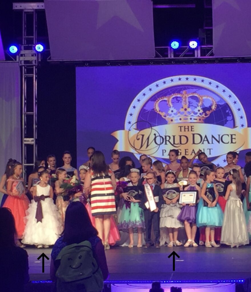 Onstage at the World Dance Pageant, a group of young dancers in formal dresses stand in several lines as one woman passes out awards.