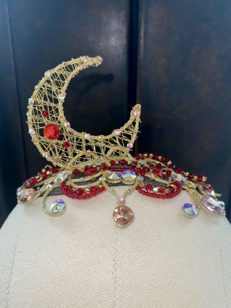 A ballet costume headpiece with red and gold gems and a gold waning crescent moon made of gold-wrapped wire.