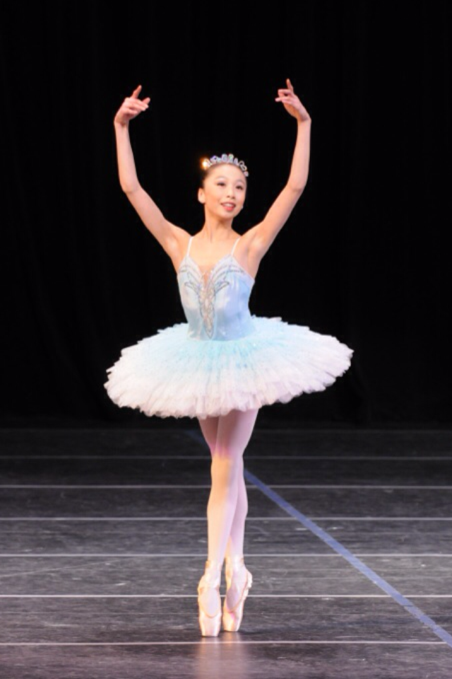 A photo of a 13-year-old Candy Tong wearing a light blue pancake tutu and tiara. She poses in sous-sus on pointe with her arms in a wide fifth position en haut.