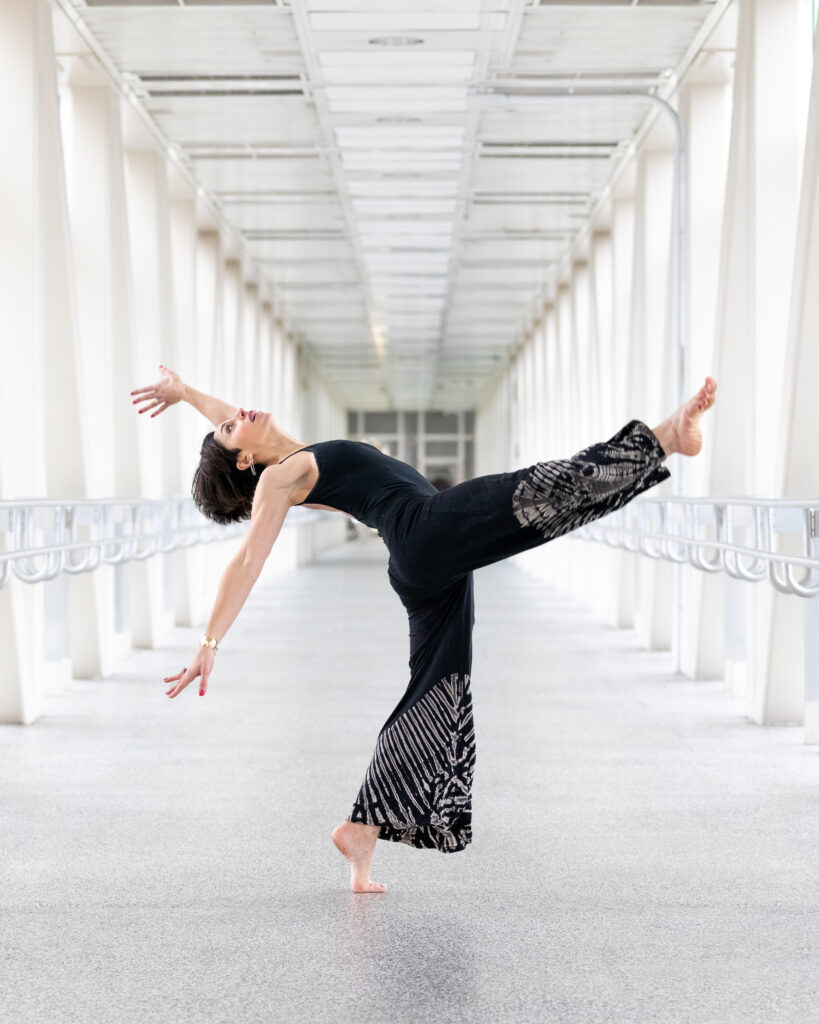 In a long, brightly lit hallway with white and light silver coloring, Julia Basso poses in profile with one leg lifted front. Her supporting leg bends slightly, on forced arch, and she leans backward with her torso so she looks up at the ceiling. Both arms reach behind her, the farthest raised above her head while the closest reaches toward the floor. She wears a black jumpsuit with white tie-dye at the bottom.