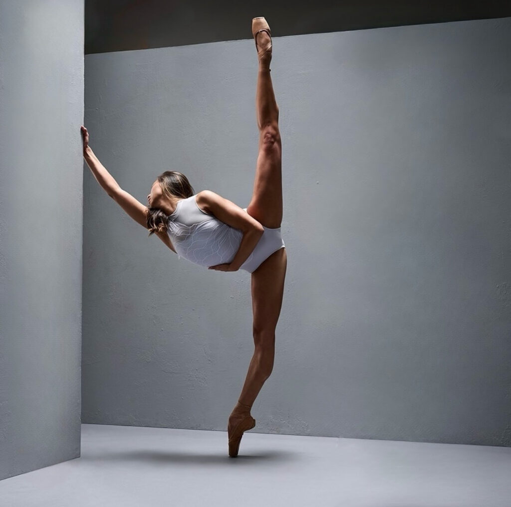 Candy Tong poses in penché en pointe with her torso twisted front. She wraps one arm around her waist while holding onto a wall with her other hand, her head turned backward to look over her extended arm. She wears a white mock-neck leotard and poses among two off-white walls in front of a black backdrop.