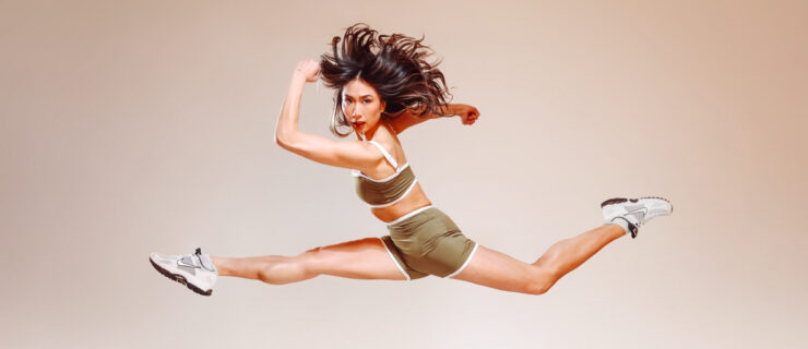 In front of a cream-colored backdrop, Candy Tong poses mid-air in a split leap with her back leg slightly bent and her arms pumping as if running. Her long hair flies around her head and she looks off-center with a determined expression. She wears an olive-colored athletic set and white tennis shoes.