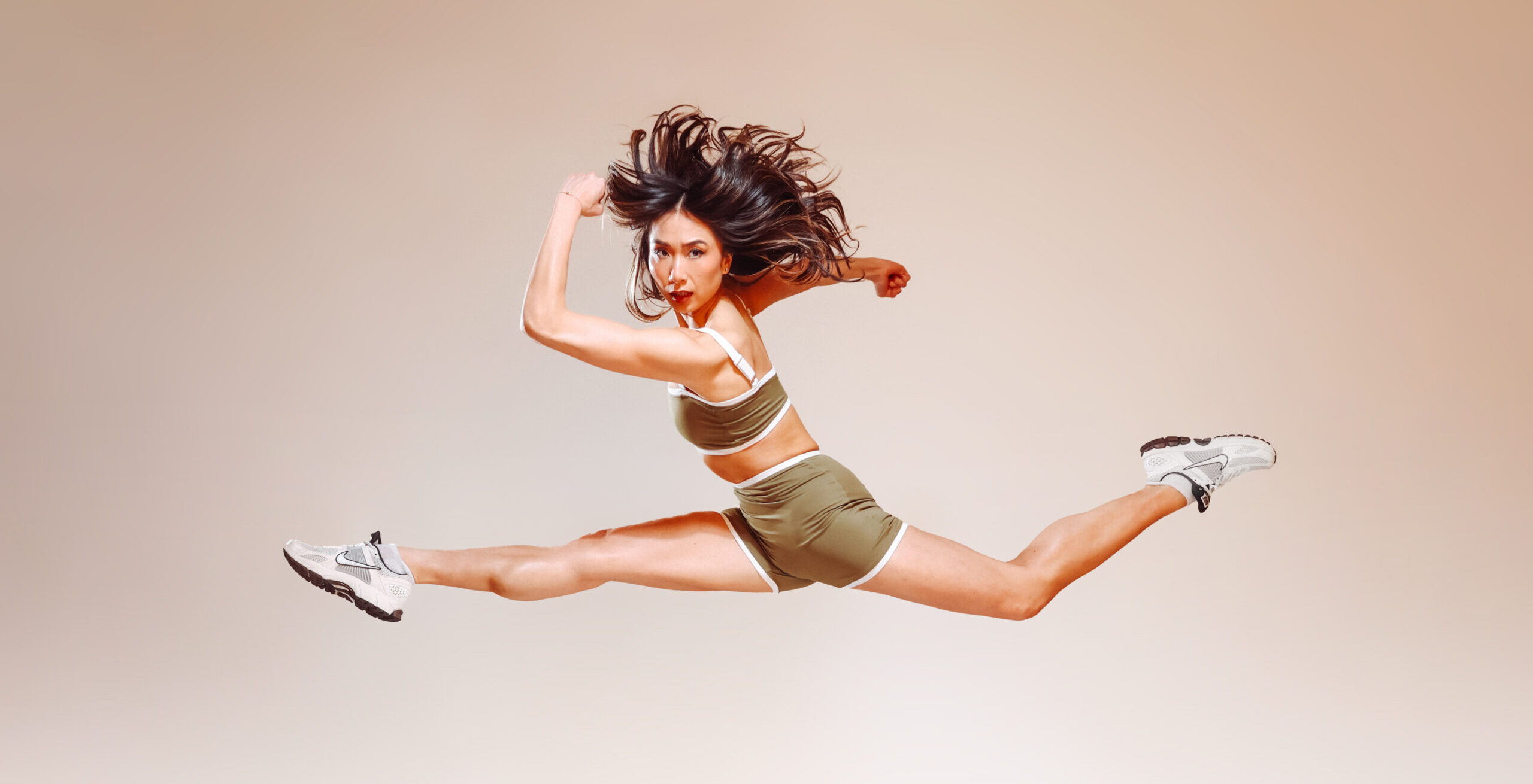 In front of a cream-colored backdrop, Candy Tong poses mid-air in a split leap with her back leg slightly bent and her arms pumping as if running. Her long hair flies around her head and she looks off-center with a determined expression. She wears an olive-colored athletic set and white tennis shoes.