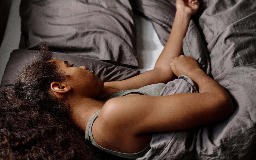 A teenage girl with thick, curly hair lies on her side in bed, with an arm on top of the gray covers.