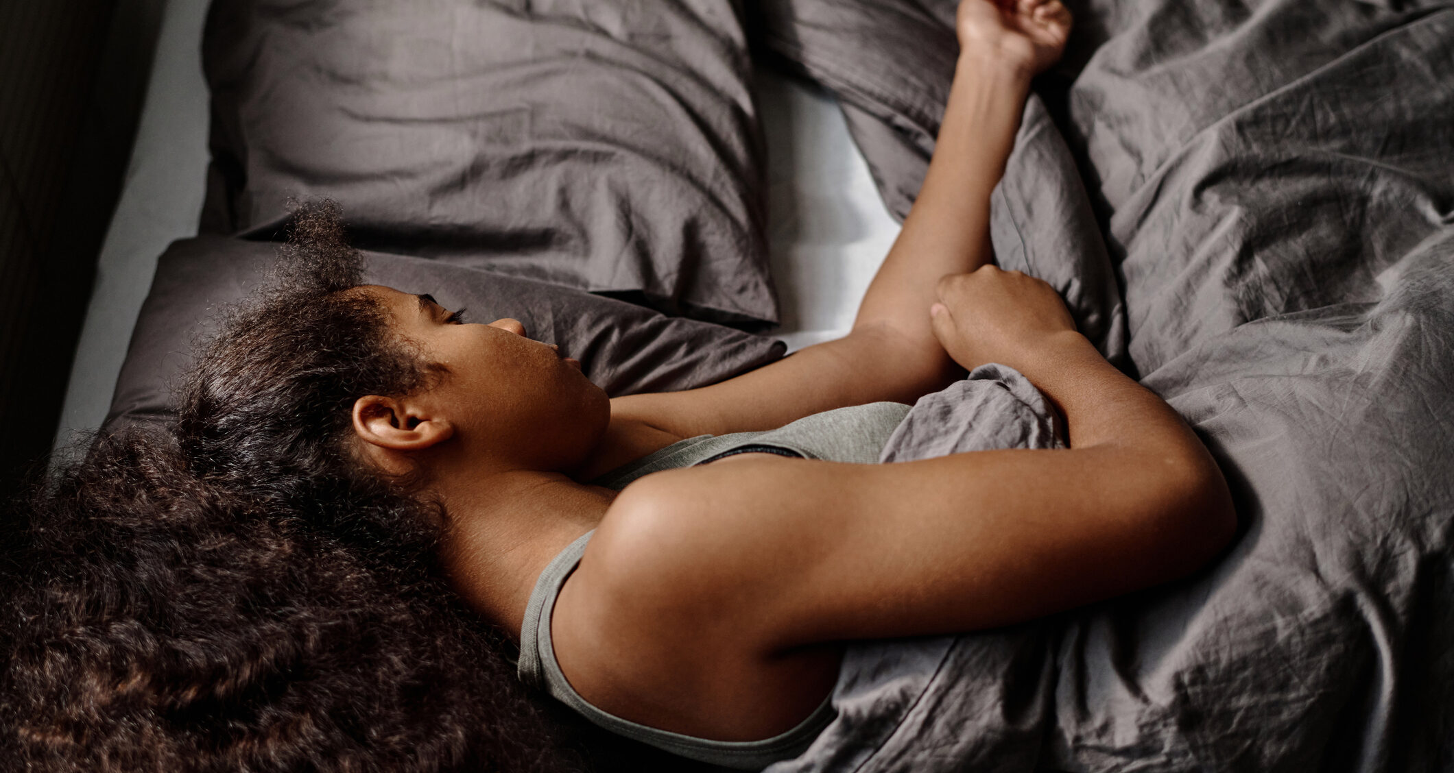 A teenage girl with thick, curly hair lies on her side in bed, with an arm on top of the gray covers.