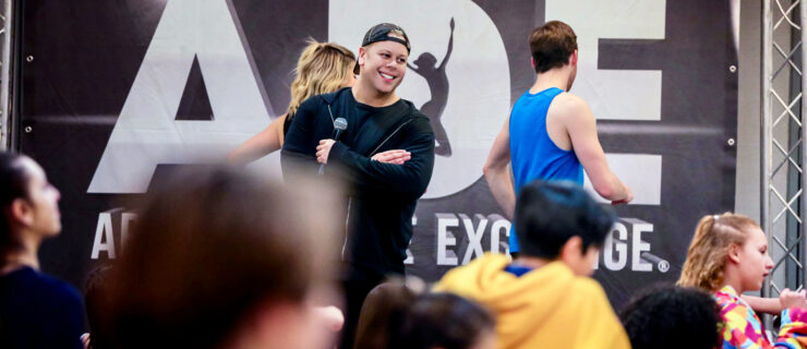 Tom Richardson stands onstage with a microphone. He smiles while watching dancers in class.