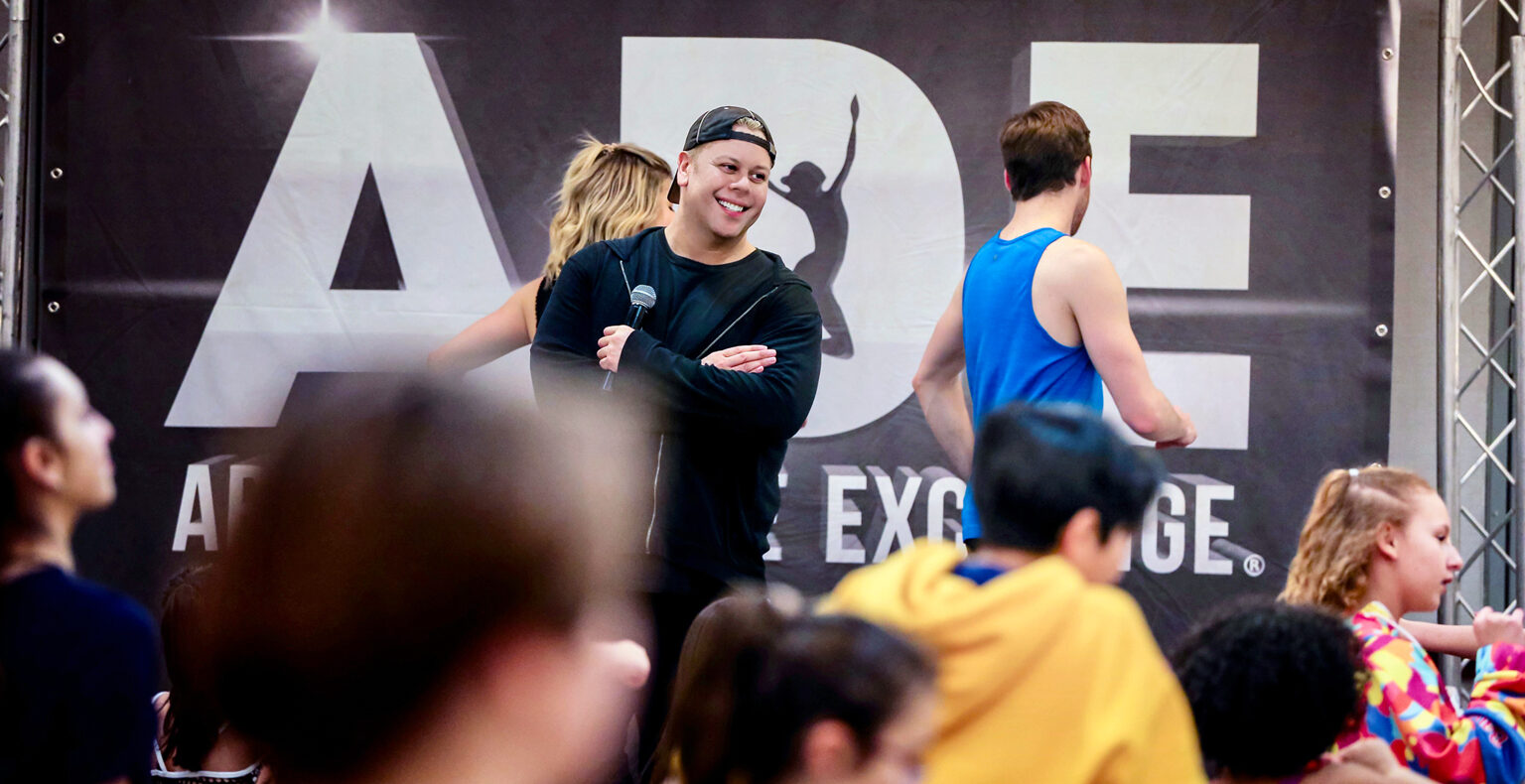 Tom Richardson stands onstage with a microphone. He smiles while watching dancers in class.