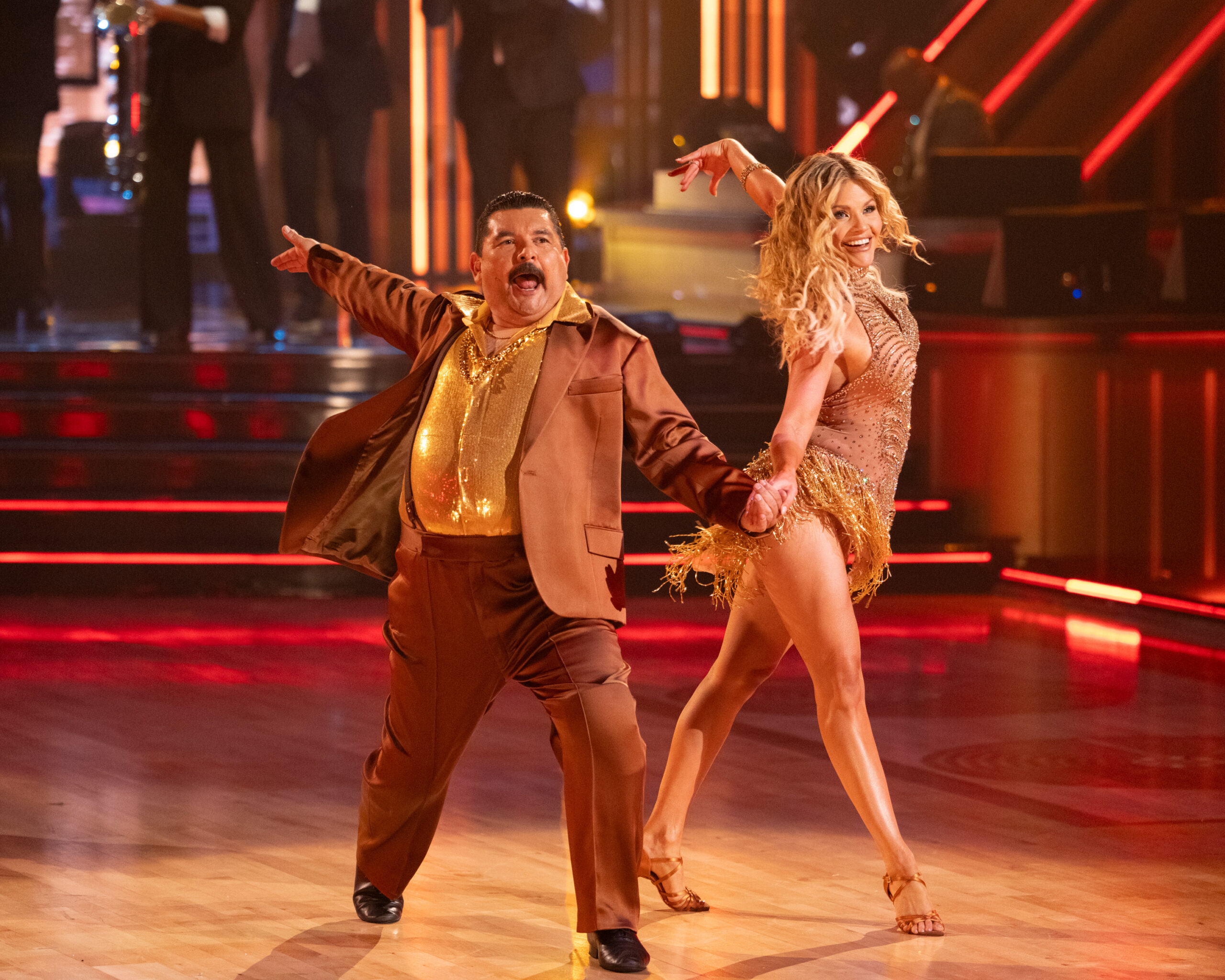 Guillermo Rodriguez and Witney Carson hold hands to the front, their other arms extended back as they twist their torsos away from each other. They each step forward with their inside legs and smile. Witney wears a sparkly gold dress with heels, and Guillermo wears a bronze tuxedo with a shiny gold shirt underneath.