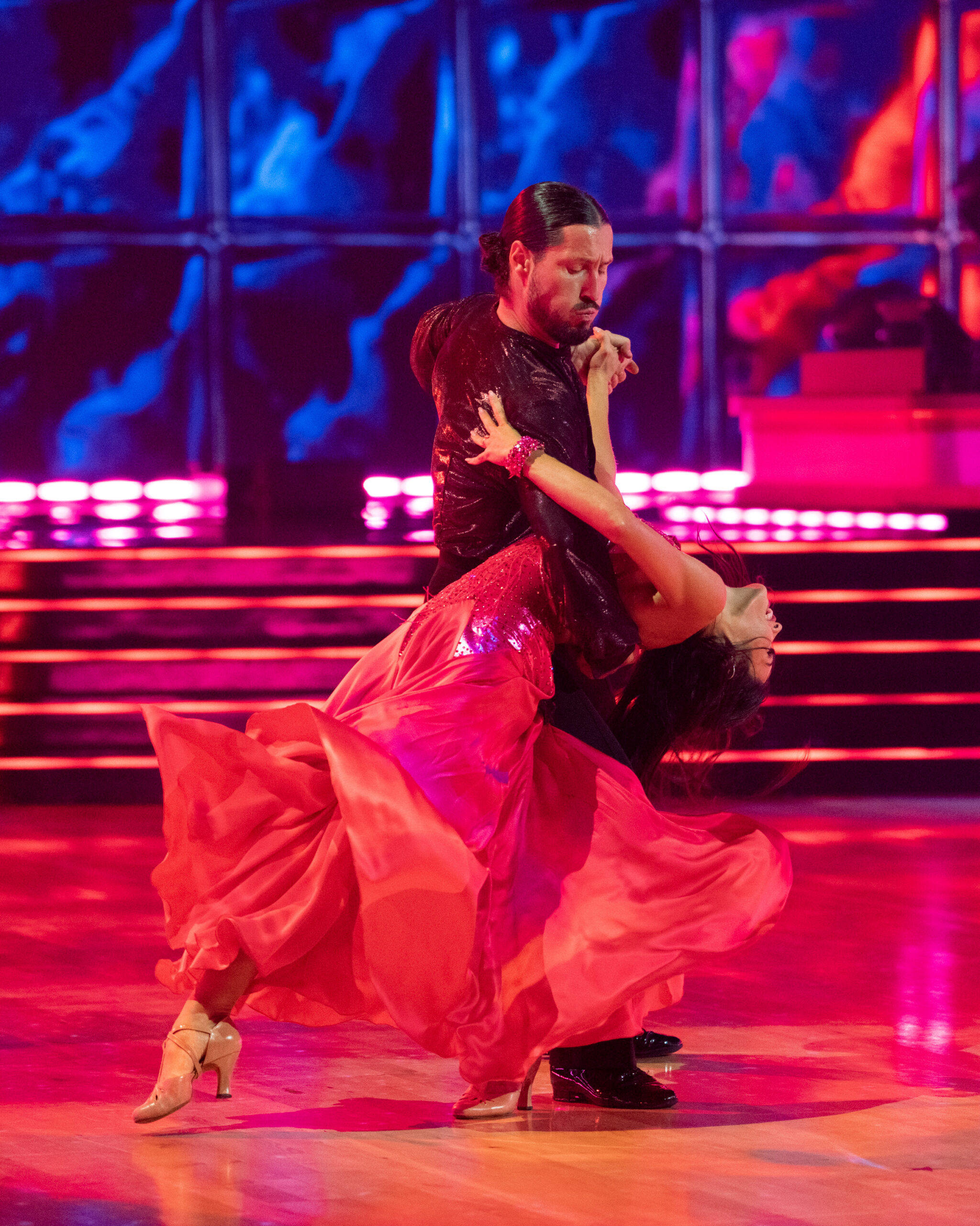 Valentin Chmerkovskiy dips Jenna Dewan as they dance together, Dewan extending her inside leg forward as she arches backward. She wears a long red dress, and Chmerkovskiy wears a shiny dark purple shirt with black pants.