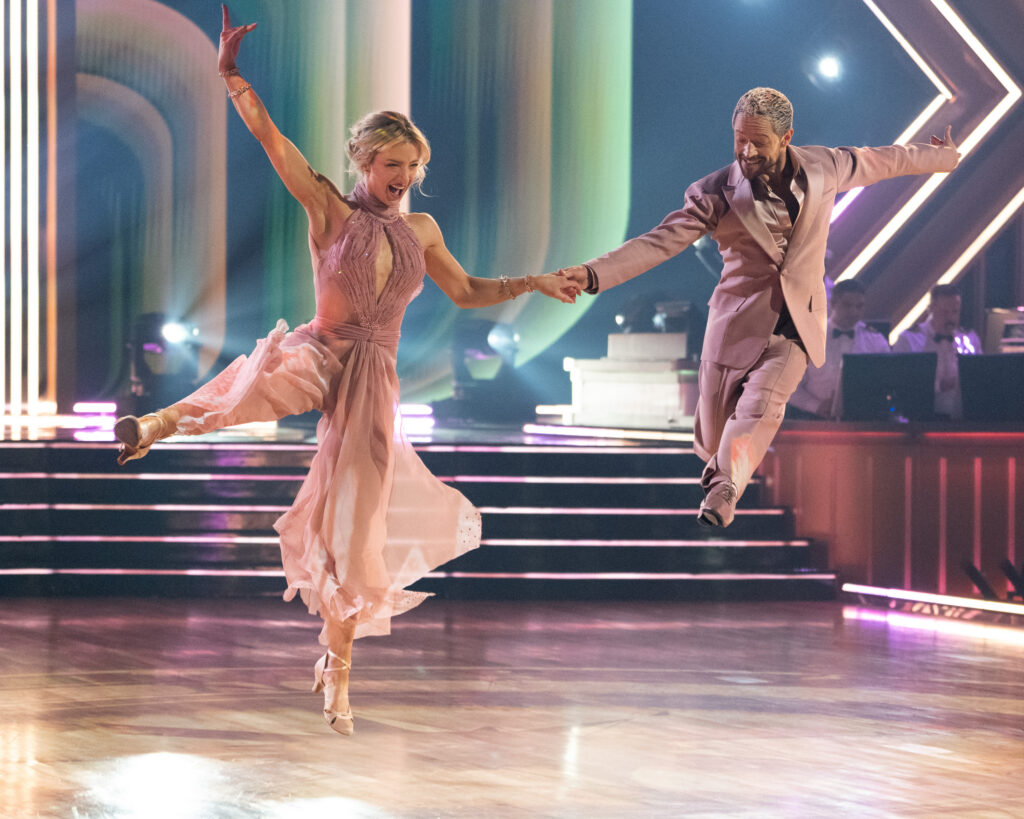 Amber Glenn and Pasha Pashkov hold hands and jump together, Glenn with one leg to the side and an arm up, and Pashkov with one leg tucked back and the other extended front.