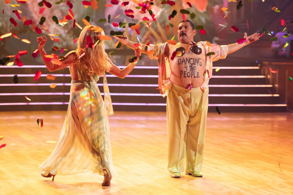 Witney Carson and Guillermo Rodriguez perform onstage as confetti drops. Carson walks with her back to the camera as Rodriguez stands with open arms, his shirt opened to reveal a coconut bra and "dancing 4 the people" written across his stomach.