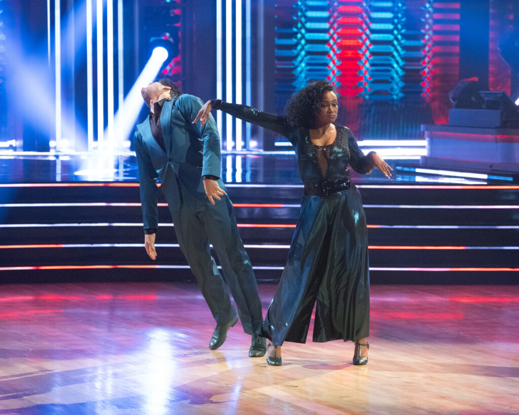 Tatyana Ali extended one arm away, hand flexed down, and turns with a satisfied expression as Jan Ravnik pretends to fall to the floor.