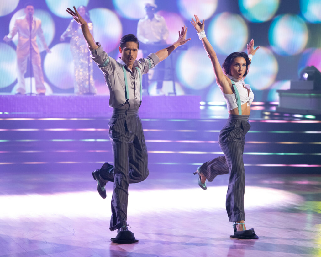 Harry Shum Jr. and Jenna Johnson, in pinstripe pants and suspenders, tuck one leg back as they step and slide forward with their bottom feet on fedoras. They make an "oh" expression with their mouths and open their arms wide in a high "V."