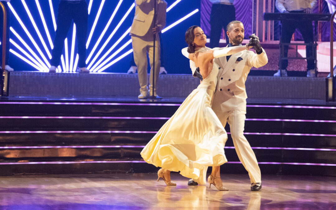 Maura Higgins and Mark Ballas dance in frame, stepping to the side with their downstage legs, as a band performs behind them.