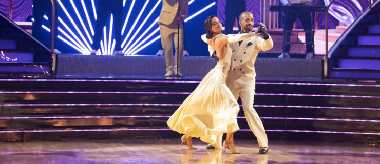 Maura Higgins and Mark Ballas dance in frame, stepping to the side with their downstage legs, as a band performs behind them.
