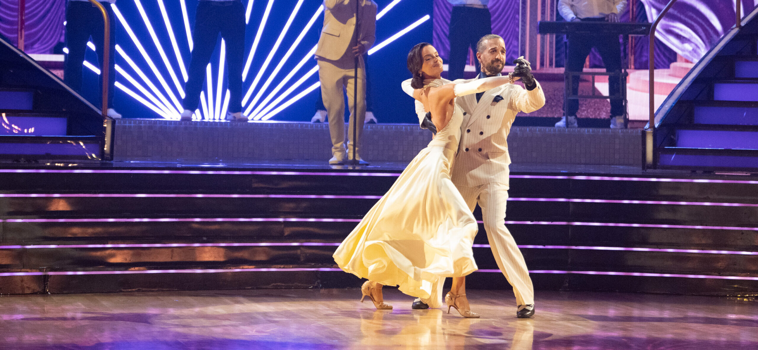 Maura Higgins and Mark Ballas dance in frame, stepping to the side with their downstage legs, as a band performs behind them.
