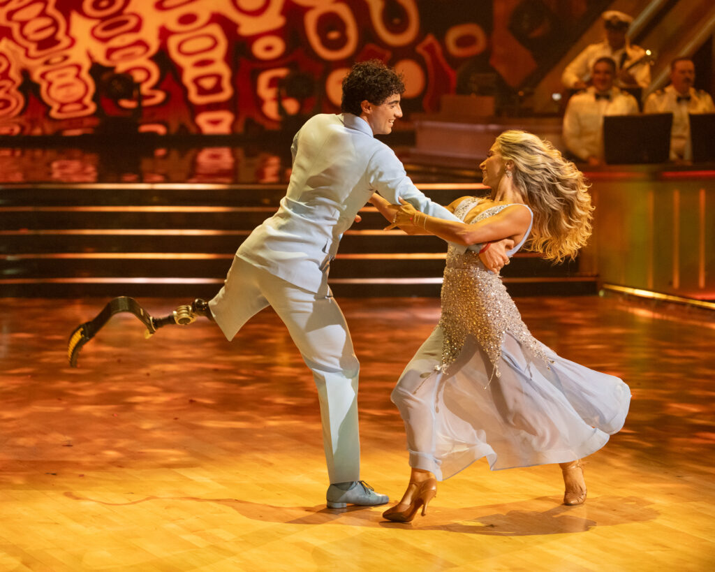 Ezra Frech and Daniella Karagach hold arms and face each other while dancing onstage in light mint-colored costumes. Frech extends one leg back in an arabesque as Karagach walks around him for a partnered promenade.