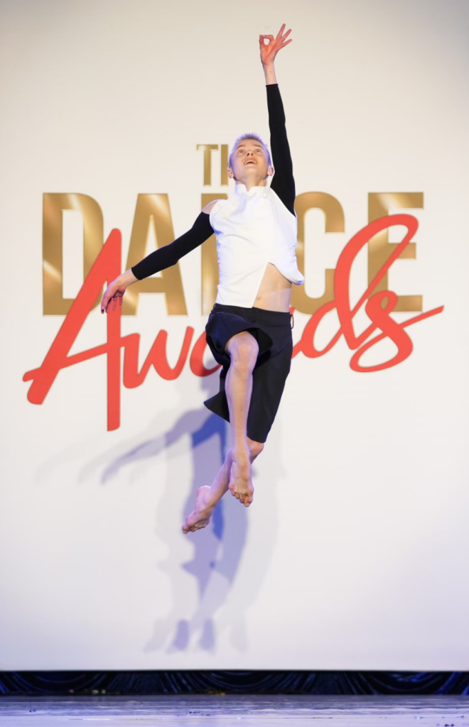 Ian Stegeman jumps with both legs slightly bent, reaching up to the sky with one arm, pointer finger and thumb touching. He looks up at that hand while his other arm trails down to the side. Behind him is a "Dance Awards" sign.