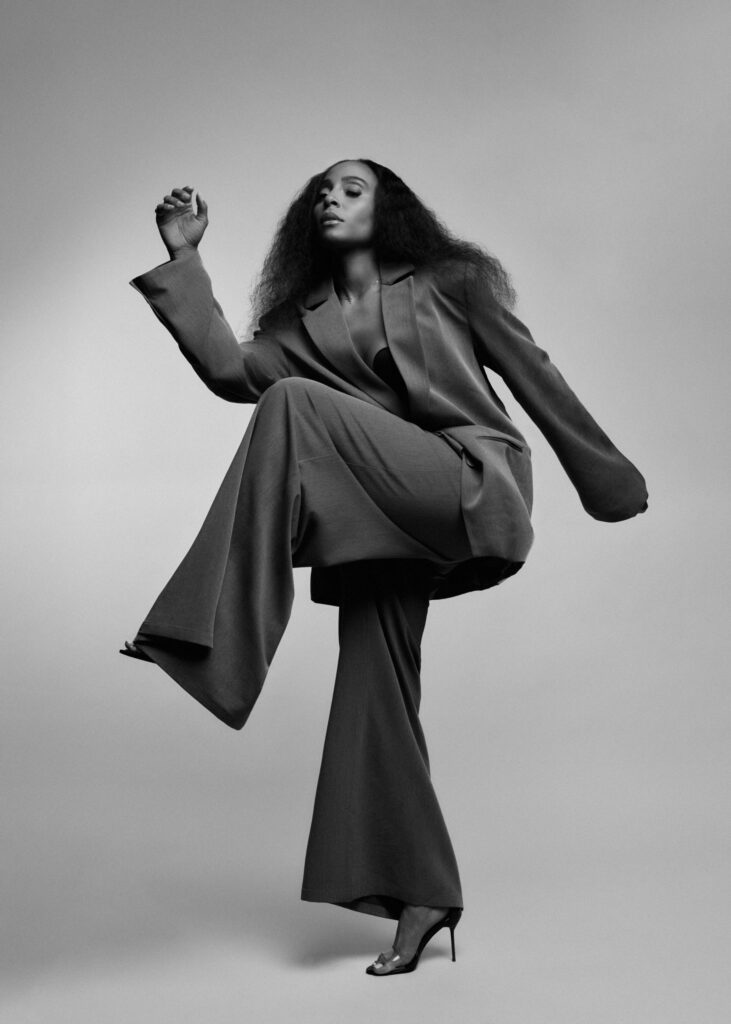 A black-and-white photo of Jasmine "JB" Badie. Badie, wearing an oversized suit, twists her hips while lifting her left leg, knee bent, across her body. She pumps one arm up gently, the other extended long, and she lifts her chin as she looks down toward the camera.