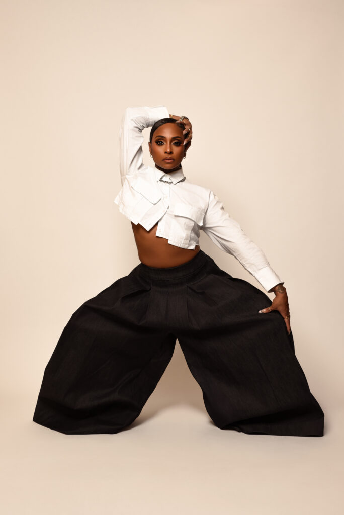 Jasmine "JB" Badie poses in front of a peach-colored backdrop wearing black wide-legged pants and a cropped white button-down. She stands with her legs wide, knees bent, and rests her left hand on her left knee with her right arm bent up and over her head, her hand resting on her opposite cheek.