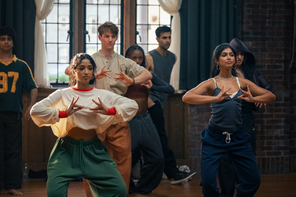 A diverse group of young adults practicing dance in a studio with wooden floors, brick walls, and large windows. The two women in the front are focused on their hand movements, suggesting a choreographed routine.
