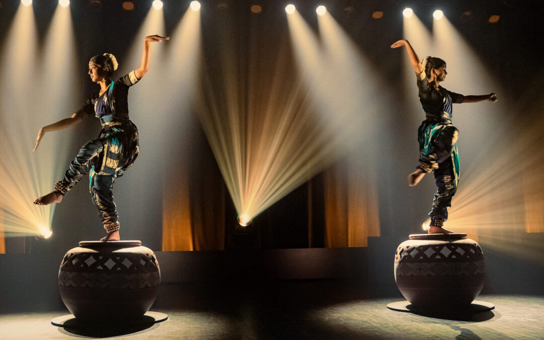 Two performers in traditional attire balancing on large decorated drums on a stage, dramatic spotlights and beams illuminating them in a dark theater setting.