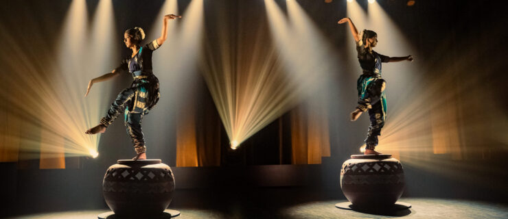 Two performers in traditional attire balancing on large decorated drums on a stage, dramatic spotlights and beams illuminating them in a dark theater setting.