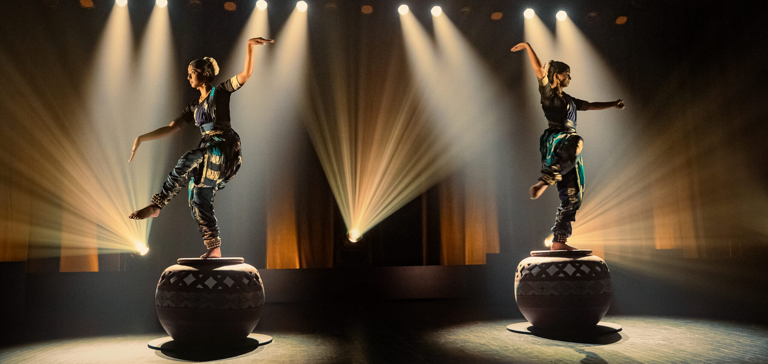 Two performers in traditional attire balancing on large decorated drums on a stage, dramatic spotlights and beams illuminating them in a dark theater setting.