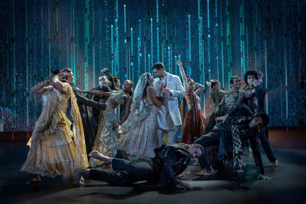 Onstage in front of a sparkly blue backdrop, a large group of dancers in traditional attire pose in a dramatic tableau.