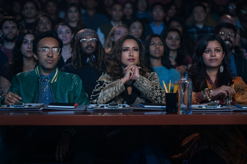 In a packed college auditorium, three judges sit at a table with clipboards and pencils. They watch a performance onstage, off-camera.