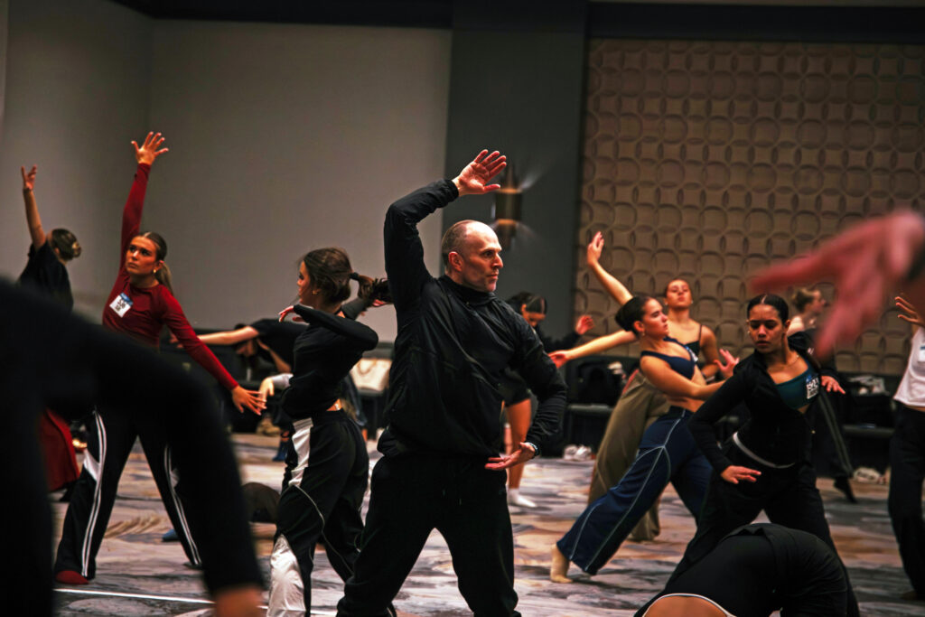 In a large hotel ballroom, Jason Parsons leads a class of teenage dancers in contemporary improv. Parsons and most of the dancers wear dark clothing.