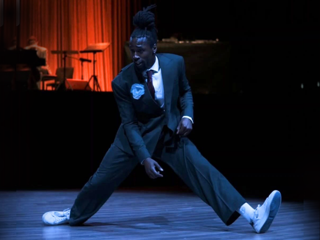 Tyedric Hill dances in a dark gray suit with a white shirt, black tie, and bright blue flower at the lapel. He splits his legs, reaching one arm down and pulling back and up gently with this other, elbow bent, with his fingers at his hip. Behind him is an elevated stage with a dark red curtain and musicians with instruments.