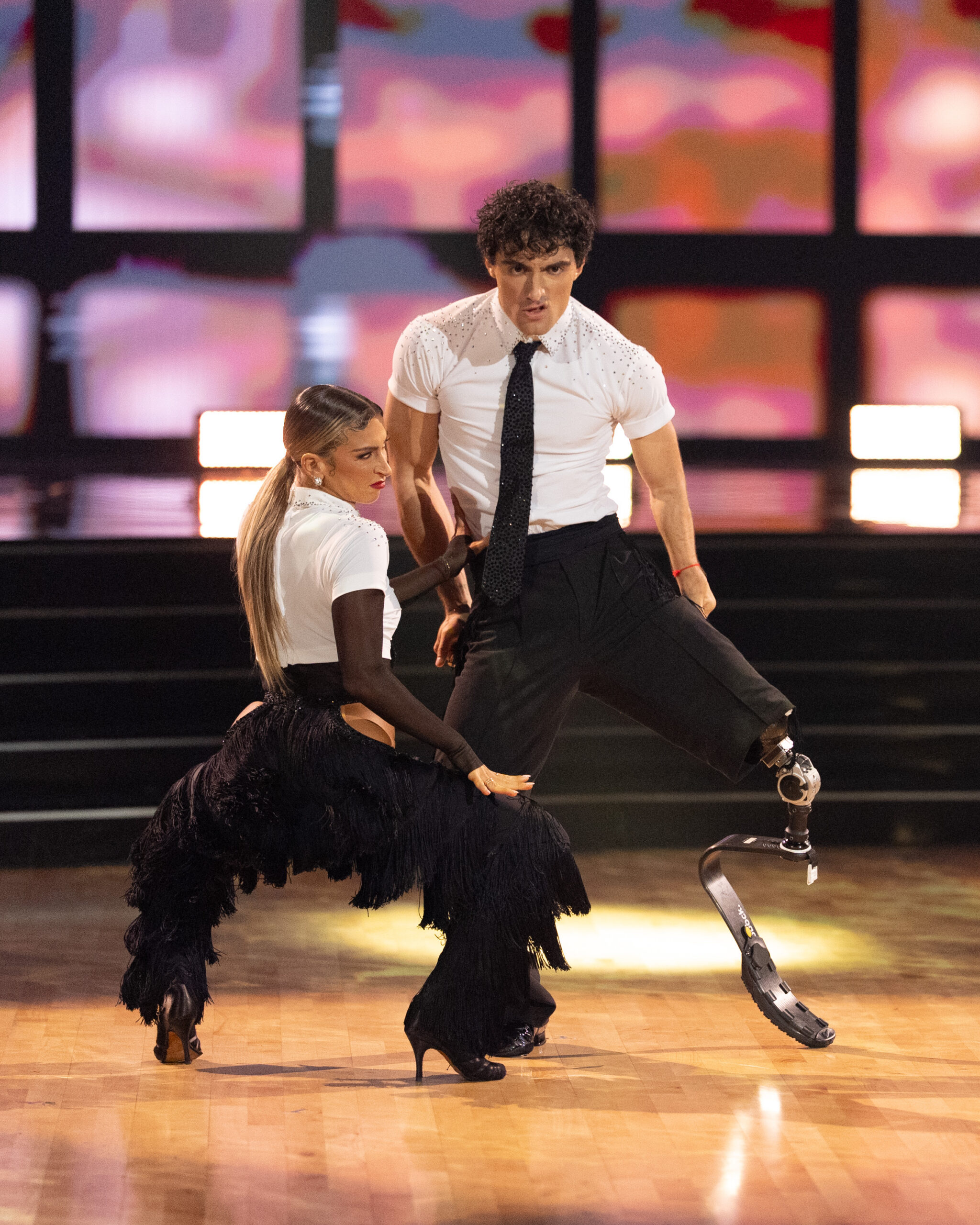Daniella Karagach, facing back, and Ezra Frech, facing forward, bend both legs while swinging their hips and torsos. They wear black pants and white tops, and both look toward the front with intense expressions.