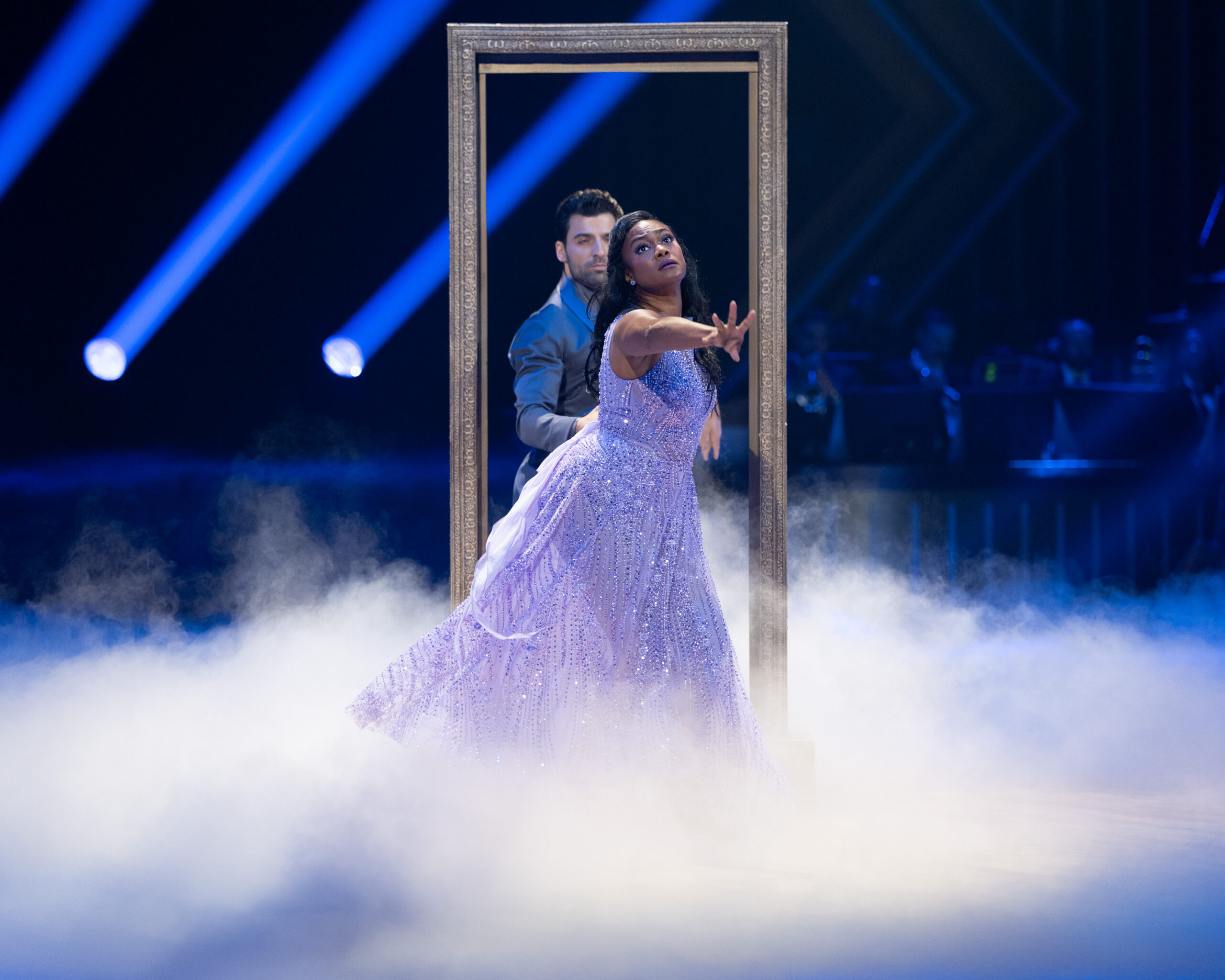Onstage, amid dry-ice fog, Jan Ravnik and Tatyana Ali dance in front of a gilded mirror frame. Ravnik stands behind Ali as she turns toward the front, her purple dress swishing, and reaches forward with a longing expression.