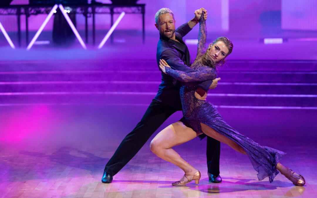 Bathed in purple lighting, Pasha Pashkov and Amber Glenn tango on the "DWTS" dance floor. Glenn lunges with her back leg extended, twisting her torso toward the camera as Pashkov mirrors her with a shallower lunge.