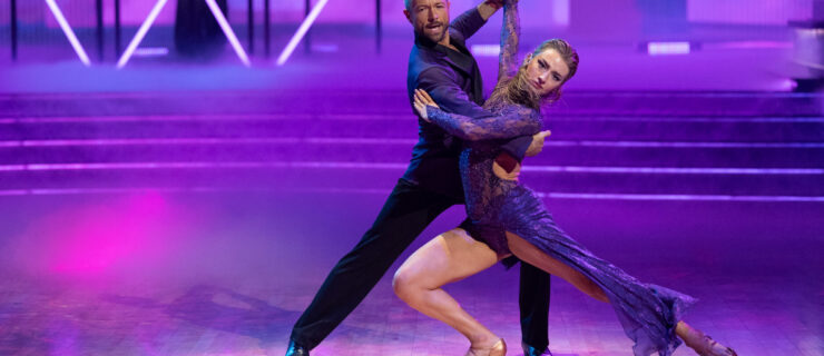 Bathed in purple lighting, Pasha Pashkov and Amber Glenn tango on the "DWTS" dance floor. Glenn lunges with her back leg extended, twisting her torso toward the camera as Pashkov mirrors her with a shallower lunge.