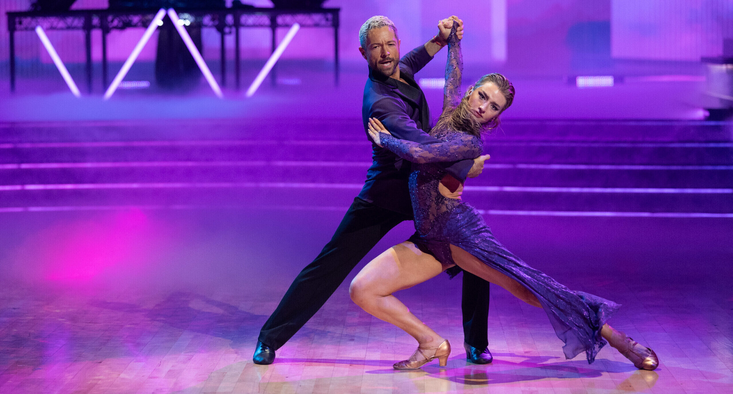 Bathed in purple lighting, Pasha Pashkov and Amber Glenn tango on the "DWTS" dance floor. Glenn lunges with her back leg extended, twisting her torso toward the camera as Pashkov mirrors her with a shallower lunge.