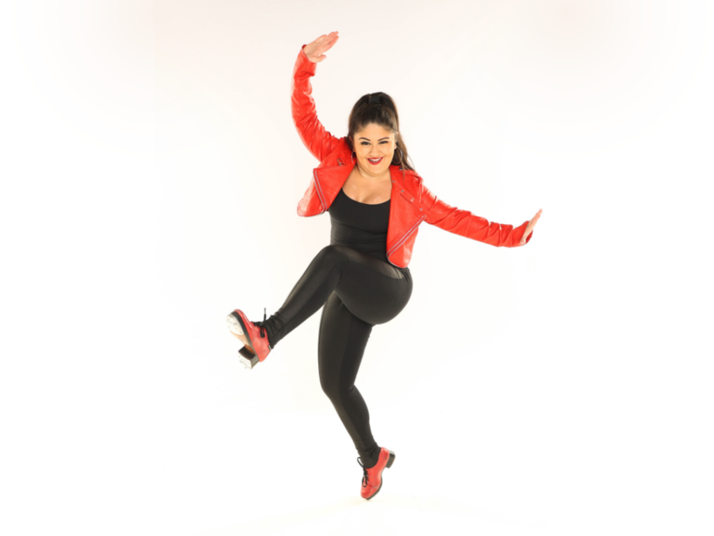 Kelsey Rose bends both legs while balancing on the tip of one tap shoe, kicking her other leg forward with a flexed foot. She lifts one arm up and one to the side with flexed hands, wearing a black bodysuit, a bright red jacket, and red tap shoes.