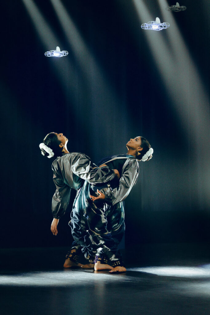 On a dark stage with two square-shaped spotlights, two dancers in traditional attire stand next to each other, facing opposite directions, and link arms while bending backward.