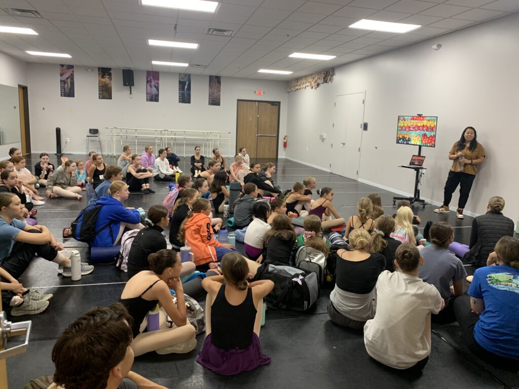In a dance studio, a large group of young dancers sit on the floor as a registered dietitian stands at the front, next to a monitor, and presents about nutrition.