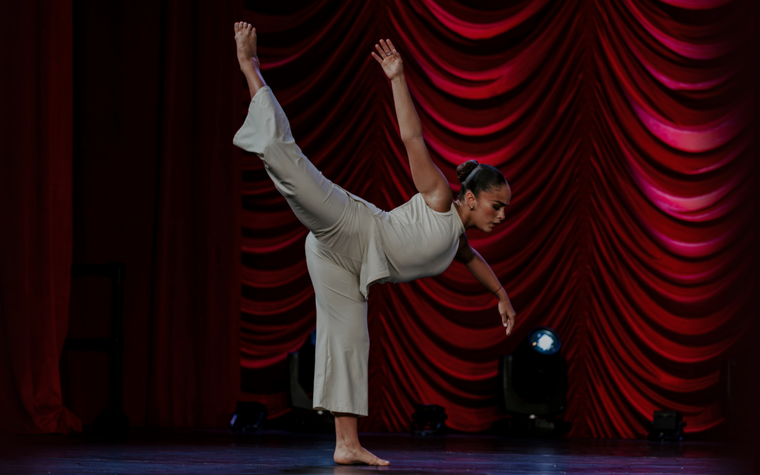 Giselle Gandarilla does an attitude penché with one arm curved to the side and the other extended up and back to follow her leg line. She wears a matching set of white pants and a sleeveless top, with her dark hair in a slicked back low bun. Behind her are draped red curtains.
