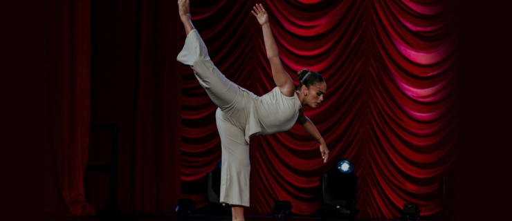 Giselle Gandarilla does an attitude penché with one arm curved to the side and the other extended up and back to follow her leg line. She wears a matching set of white pants and a sleeveless top, with her dark hair in a slicked back low bun. Behind her are draped red curtains.