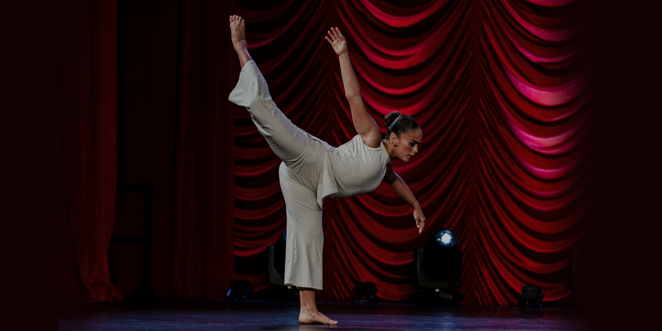 Giselle Gandarilla does an attitude penché with one arm curved to the side and the other extended up and back to follow her leg line. She wears a matching set of white pants and a sleeveless top, with her dark hair in a slicked back low bun. Behind her are draped red curtains.