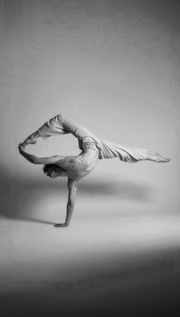 A black-and-white photo of Joey Vice balancing on one hand, his legs split horizontally with his top leg in a high attitude. He reaches up with his other hand to grab his top foot. Vice wears linen pants and is barefoot.