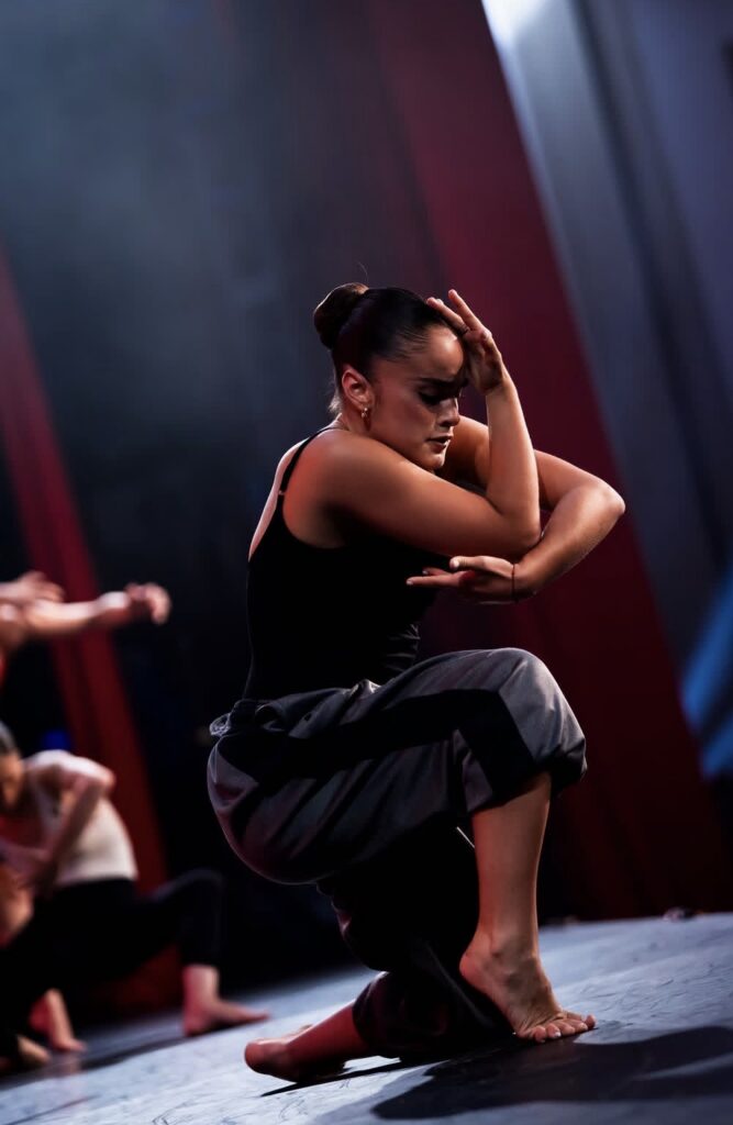 Giselle Gandarilla crouches with her front foot in forced arch, bending one elbow to the side with her palm on her knee. She bents her other elbow to rest on top of that same knee, framing her face with her hand. She wears a black leotard and dark sweatpants.
