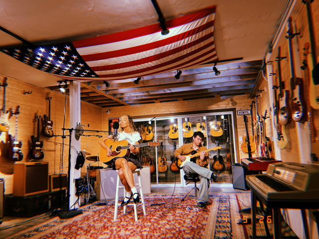In a large room with Americana decor, two siblings sit on stools and play guitars together.