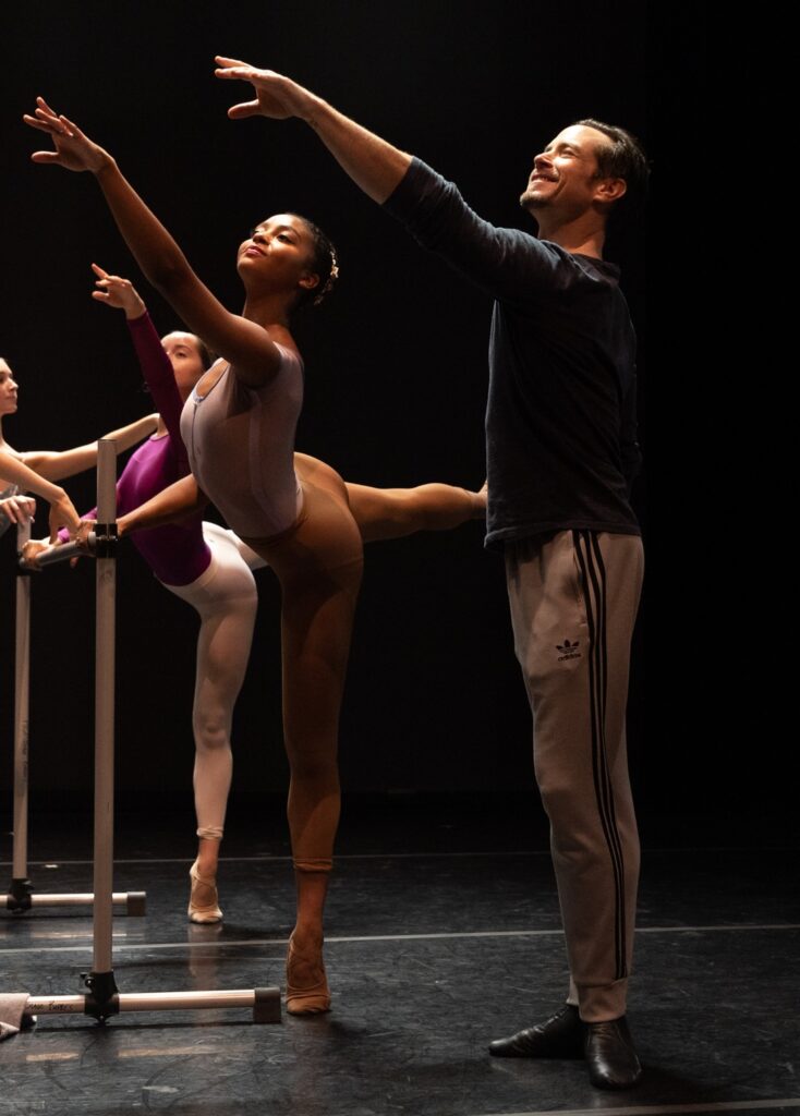 Sascha Radetsky teaches a ballet class, demonstrating an arabesque arm as Maddie Brown does an arabesque on pointe at the barre.