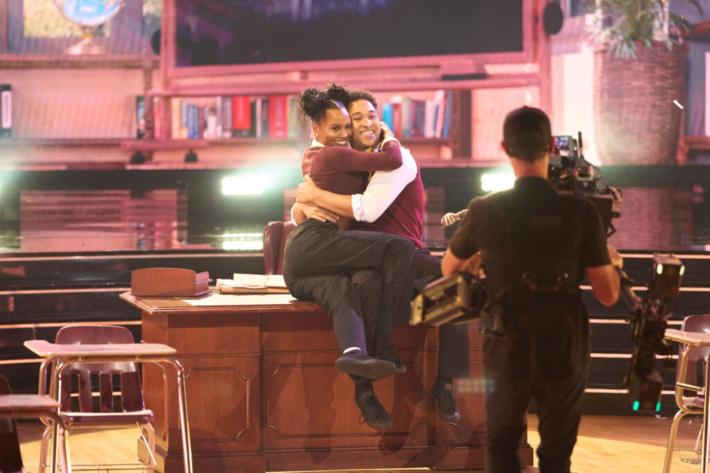 Ciara Miller and Brandon Armstrong, dressed in school uniform costumes, sit on a teacher's desk and hug while a camera man videotapes them.