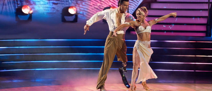 Ezra Frech and Danielle Karagach perform a rumba onstage with pink, blue, and yellow lighting. Frech places a hand on Karagach's shoulder as they step forward with their left legs and sway their hips with opposition in the torso. They extend their other arms out long with a curve.