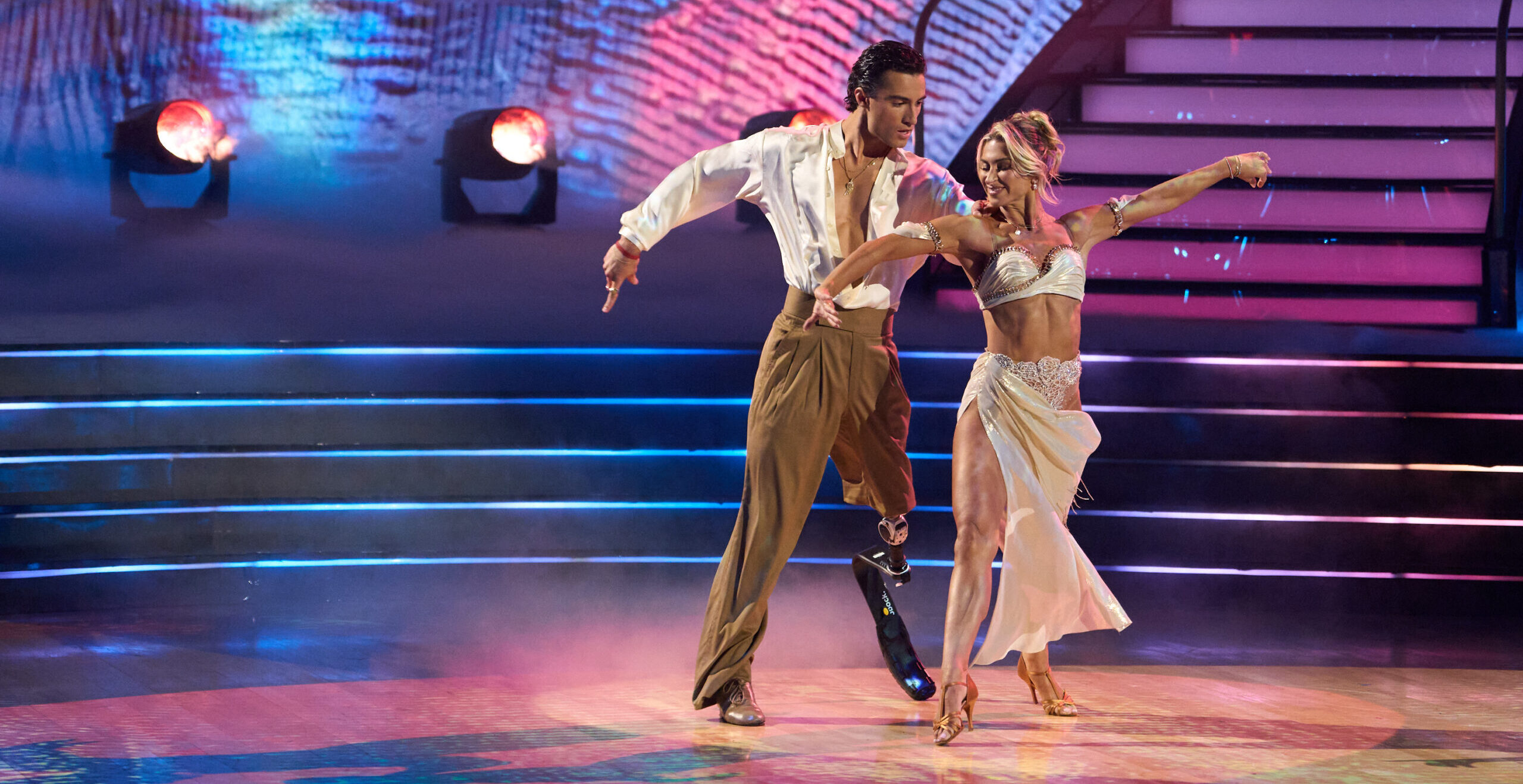 Ezra Frech and Danielle Karagach perform a rumba onstage with pink, blue, and yellow lighting. Frech places a hand on Karagach's shoulder as they step forward with their left legs and sway their hips with opposition in the torso. They extend their other arms out long with a curve.